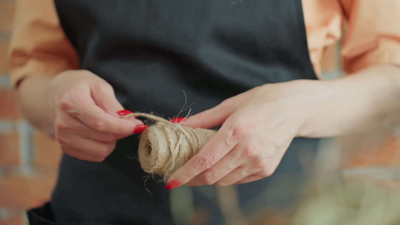 Decorator in black apron holding spool of twine, pulling natural string with both hands, preparing craft material for handmade decoration, focusing on artisan process, rustic workspace, preparation