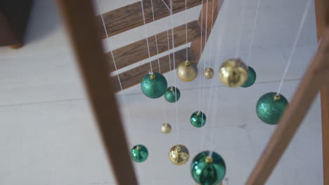 wide top shot of green and golden christmas tree decorations balls, hanging on white thread from a wooden ladder in slowmotion