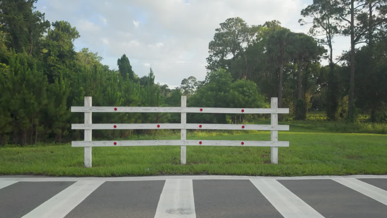 Forward motion across pavement toward a white wooden fence with trees and a cloudy sky in the background.