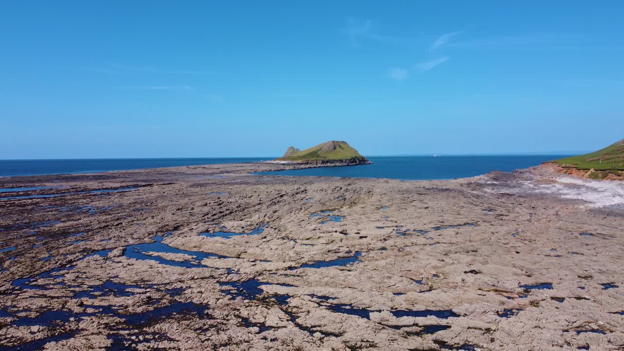 Rising Sideways Aerial Drone View of Worm's Head at Low Tide in Rhossili Bay with Dozens of Rock Pools and Calm Blue Ocean Sea. Rocky Coastline in Wales, UK.