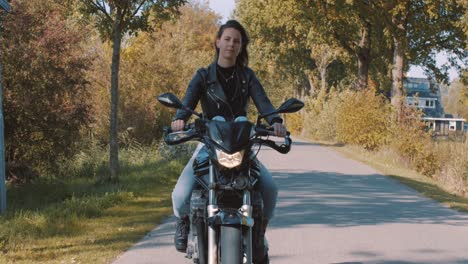 Hübsche-Lächelnde-Europäische-Junge-Frau,-Die-An-Einem-Sonnigen-Tag-Ein-Motorrad-Trägt,-Das-Lederjacke-Im-Wald-Mit-Lebhaften,-Bunten-Goldenen-Herbstblättern-Trägt