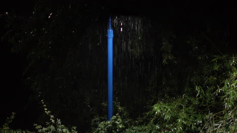 la lluvia torrencial de verano cae más allá de la luz emitida por un poste de luz azul rodeado de follaje verde por la noche mientras los insectos bailan alrededor de la fuente de luz