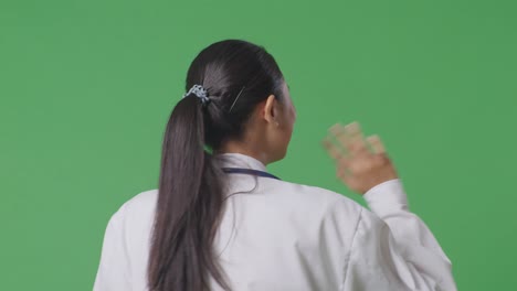 vista de atrás de una mujer asiática con un estetoscopio agitando la mano y sonriendo mientras camina en el fondo de la pantalla verde en el hospital