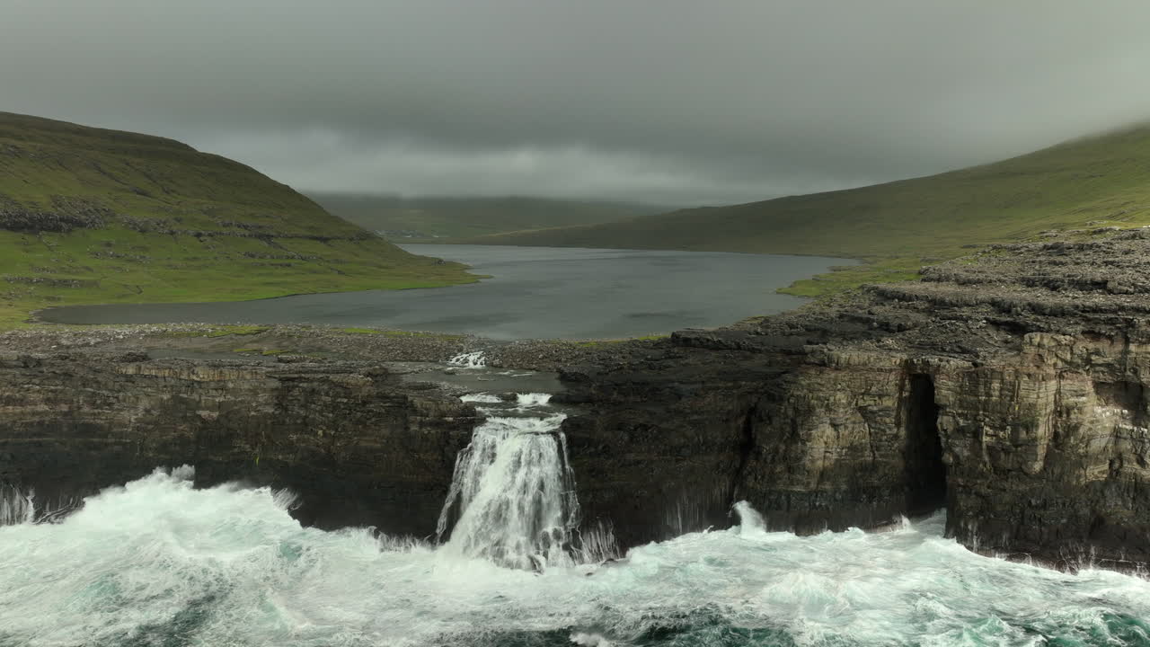 Lake Sorvágsvatn pours its waters into sea on cloudy day, Faroe Islands. Aerial backwards