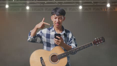 asian teenager with guitar pointing to smartphone in hand on stage