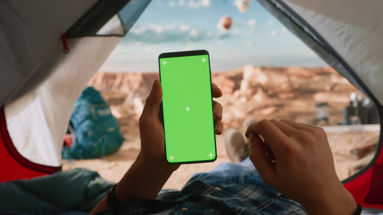 vista de un turista sosteniendo verticalmente un teléfono inteligente con un marcador de posición de pantalla verde, deslizando un dedo sobre la pantalla. viajero descansando en una tienda en la cima de una montaña rocosa y volando globos de aire caliente.