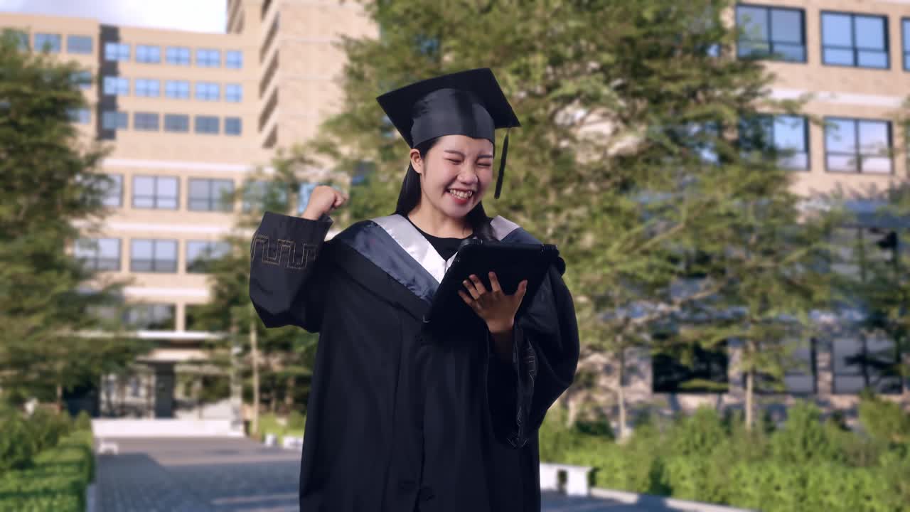 estudiante asiática se gradúa con gorra y bata mirando una tableta luego gritando gol celebrando frente a un magnífico edificio universitario