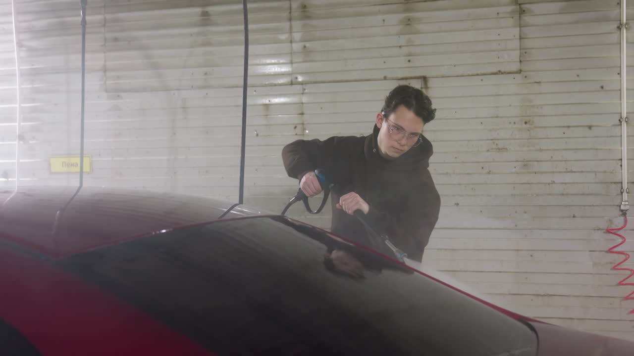 young man in hoodie and eyeglasses rinsing red car with high pressure hose inside garage under sign on wall capturing dynamic spray and foam removal with concentrated focused expression