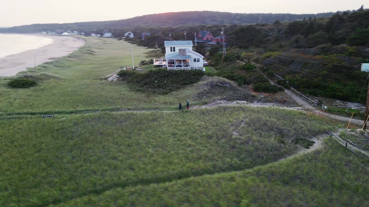 Episk luftfoto af et ungt par, der går gennem græsklædte klitter mod en naturskøn og afsidesliggende strand ved solnedgang i Popham Beach, Maine, en populær feriedestination i det nordøstlige USA.