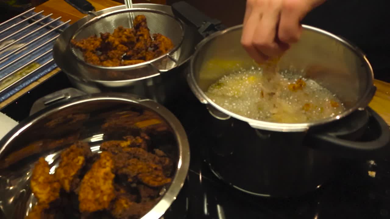 White hand placing flour coated and seasoned chicken pieces into hot yellow boiling deep frying oil in a steel gray or grey pot in a home kitchen. Ready golden brown cooked chicken on the side.
