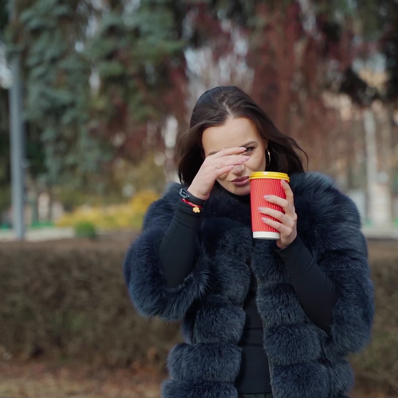 Beautiful girl drinks coffee outdoors. Portrait of a fashionable woman in fur coat with hot drink looking on camera. Stylish model in the park.