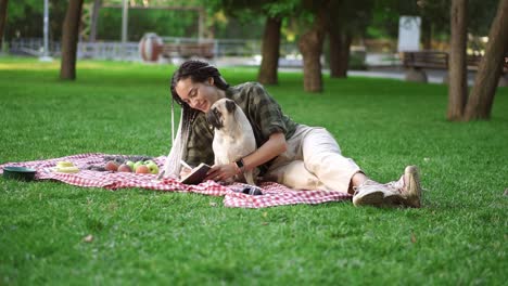 chica alegre acostada en cuadros en el césped en un parque y haciendo notas mientras un pequeño pug sentado a su lado