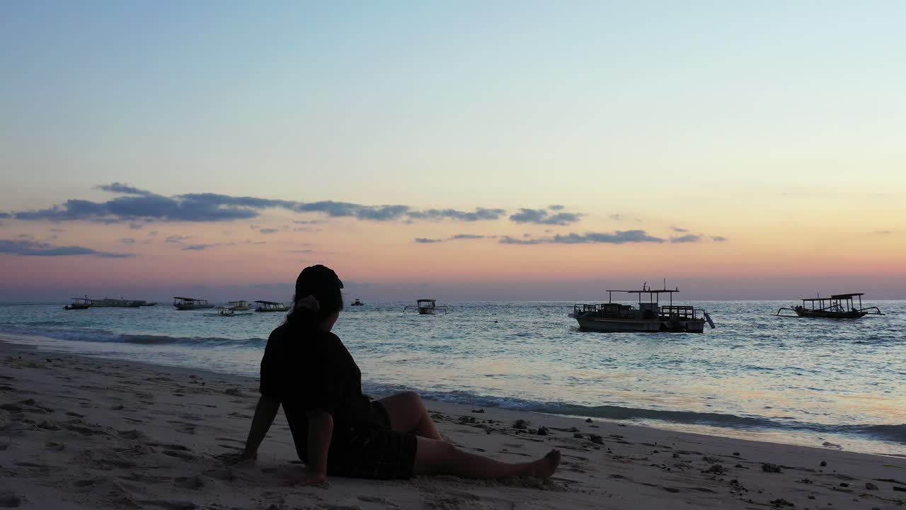 Young tourist woman with hat and t-shirt sitting on quiet exotic beach, watching beautiful twilight and anchored boats on calm lagoon in Philippines