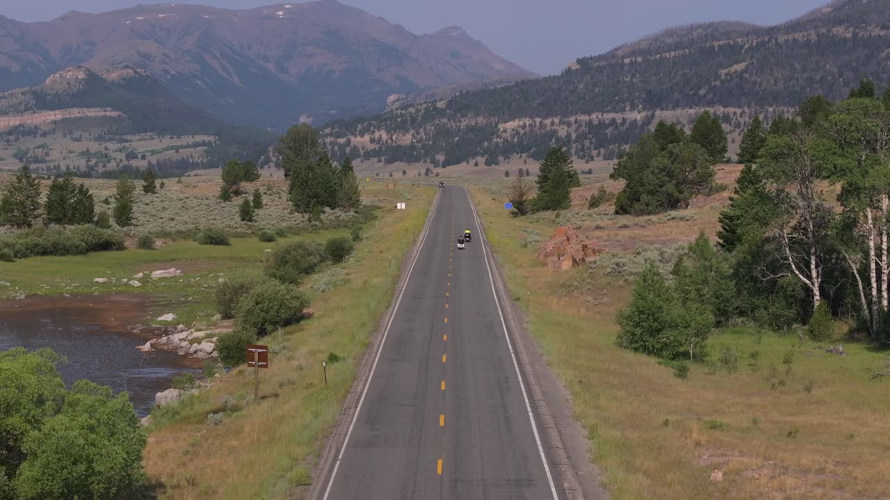 Two motorcycles ride down a scenic mountain road with lush greenery on a sunny day