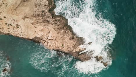 Overhead-Drohnenaufnahmen-Zeigen-Atemberaubendes-Türkisfarbenes-Wasser,-Während-Weiße-Wellen-Auf-Einen-Hellbraunen-Felsen-Vor-Der-Küste-Von-Gozo-Schlagen-Und-Die-Gischt-Hoch-Steigt