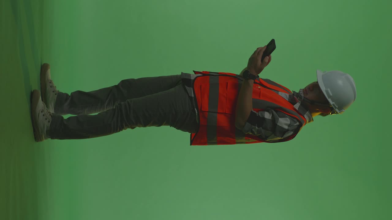 vista lateral completa del cuerpo de un ingeniero masculino asiático con casco de seguridad usando un teléfono inteligente mientras está de pie en el estudio de fondo de pantalla verde, durante la puesta o el amanecer