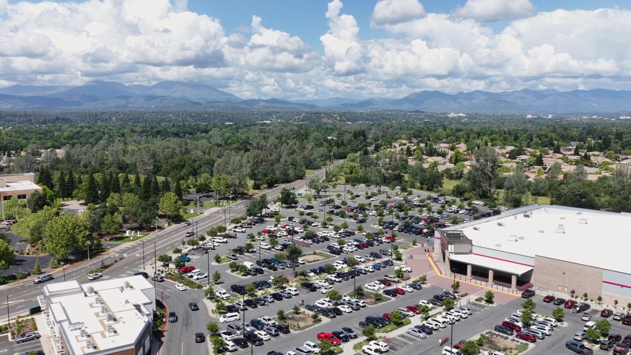 Un parcheggio affollato circonda un grande negozio al dettaglio con finiture rosse a Redding, una strada trafficata si estende oltre alberi sparsi verso distanti montagne nebbiose sotto un cielo parzialmente nuvoloso. Ampia ripresa aerea con drone