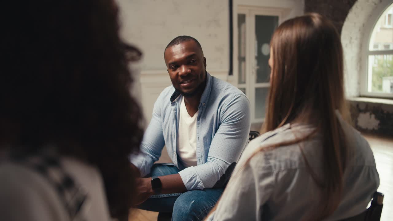 un hombre de piel negra con una camisa azul se comunica con una chica rubia en terapia de grupo en una habitación blanca. comunicación en parejas en terapia de grupo
