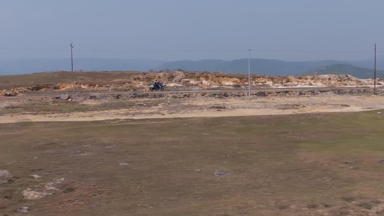 Una moto viaggia attraverso una cresta di altopiano arido con catene montuose distanti visibili nel Parco Mawdngong, Cherrapunji, Meghalaya, India. Un'ampia vista paesaggistica mostra terreno roccioso e linee elettriche