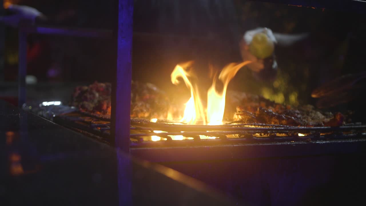 Mani guantate che girano il pollo arrosto su una griglia a fuoco vivo di notte, inquadratura ravvicinata.