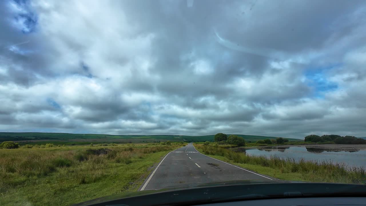POV in rapido movimento lungo una strada di campagna dritta con alberi e un lago su un paesaggio aperto e nuvole scure sopra nel Galles del Sud, Regno Unito. Pecore al pascolo a lato della strada. Clip di viaggio, trasporto, natura.