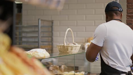 Vista-Lateral-De-Un-Hombre-De-Piel-Negra-Con-Una-Camiseta-Blanca-Y-Un-Delantal-Negro-Colocando-Productos-Horneados-En-El-Mostrador-De-Un-Supermercado-Moderno.-Un-Empleado-De-Un-Gran-Supermercado-Coloca-Productos-Horneados-En-Una-Vitrina-De-Vidrio-Especial.