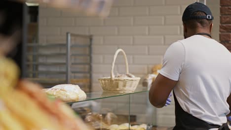 vista lateral de un hombre con color de piel negra en una camiseta blanca y un delantal negro colocando productos horneados en el mostrador en un supermercado moderno. un empleado de un gran supermercado coloca productos horneado en una vitrina especial