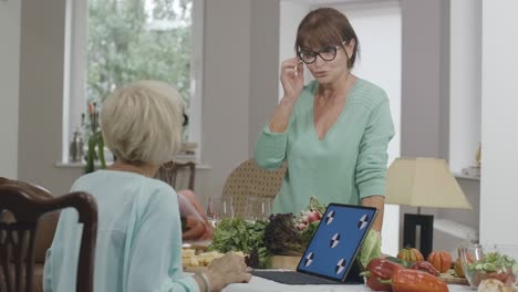 portrait of beautiful senior caucasian woman in eyeglasses talking with friend sitting at table with blue screen laptop. elegant retirees cooking and chatting indoors. cinema 4k prores hq.