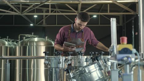 young male brewer wearing a leather apron supervise the process of beer fermentation at a modern brewery factory