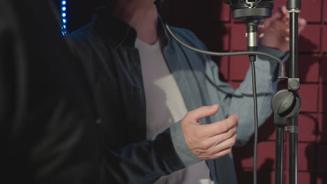 close-up of two people in a recording studio, captured mid-performance with no visible faces. one wears a blue shirt and white undershirt, moving hands expressively as they sing into a microphone