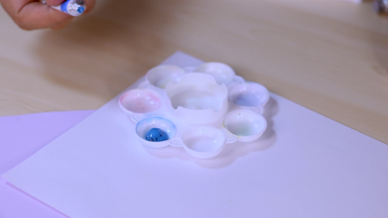 Hand mixes blue paint with brush in white palette on desk, soft lighting, close-up view