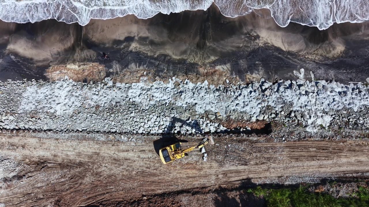 Construction work near the shore shows a yellow machine placing rocks along the coastline while waves crash at the edge during daylight