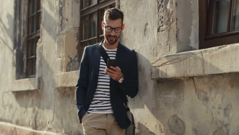 Smiling-Young-Man-In-Stylish-Outfit-Leaning-Against-The-Wall-In-The-Street,-Tapping-And-Scrolling-While-Chatting-On-The-Smartphone