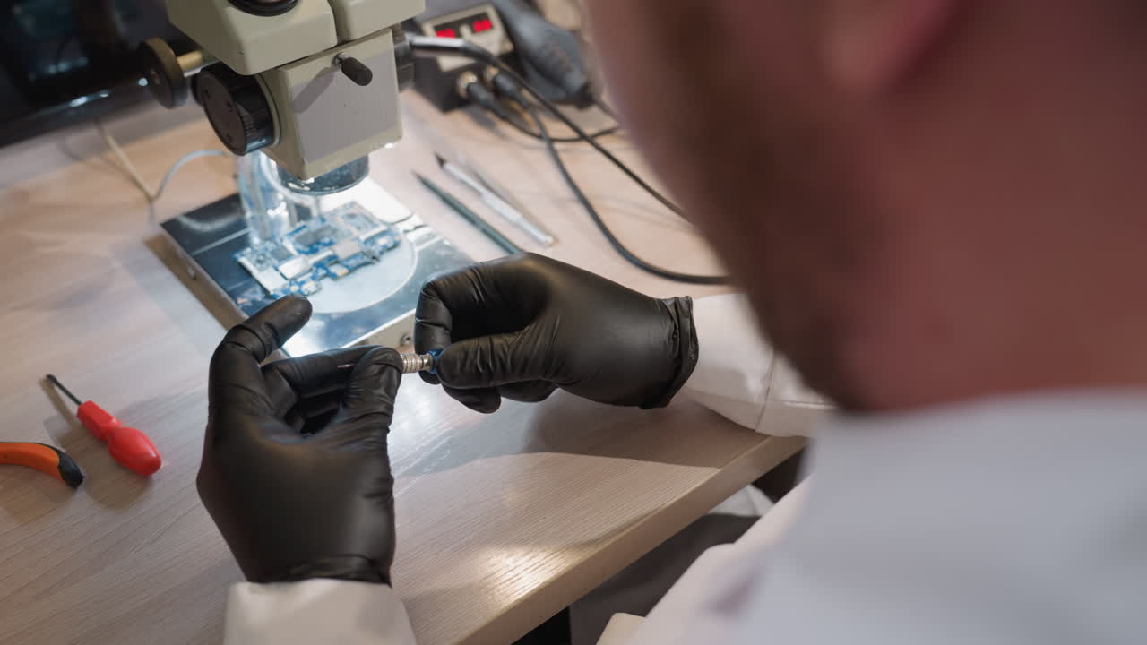 微鏡の下で回路板に取り組んでいる技術者黒い手袋をかぶった技術者は手でスクリューを回しています机の上には様々なツールが見えます