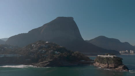 Sideways-aerial-pan-showing-the-Joatinga-beach-on-a-hazy-backlit-day-with-the-Gavea-mountain-behind-and-an-island-with-recreational-construction-on-top-in-the-foreground
