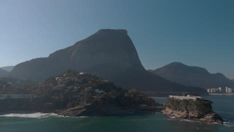 paneo aéreo lateral que muestra la playa de joatinga en un día brumoso retroiluminado con la montaña givea detrás y una isla con una construcción recreativa en la parte superior en primer plano