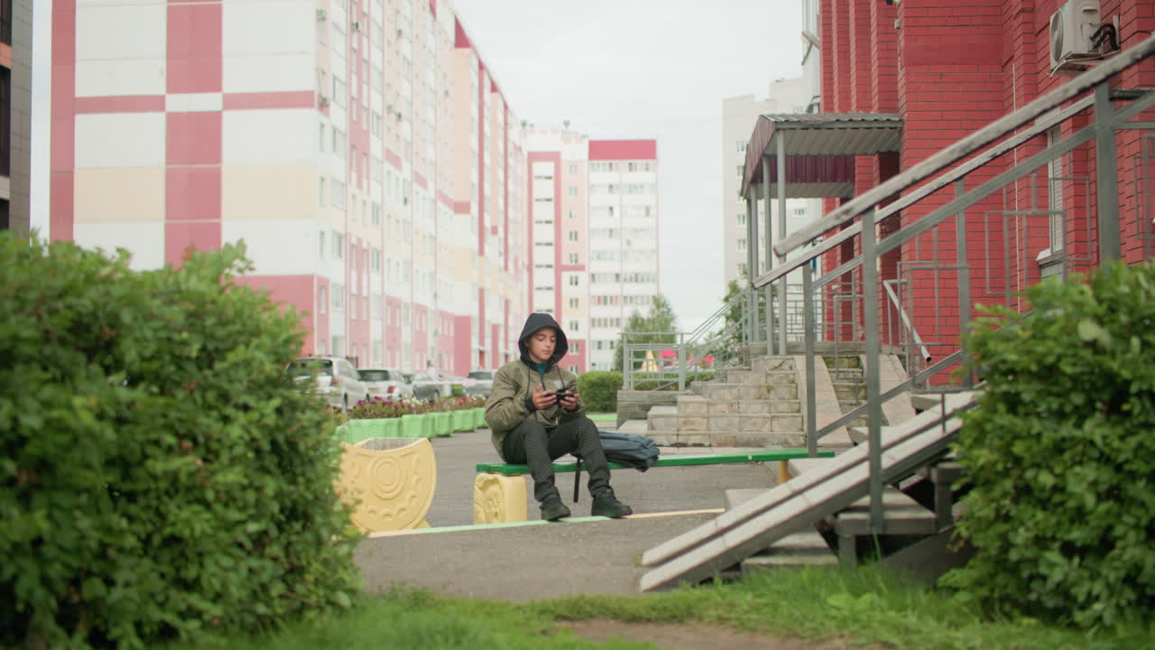Kid seated alone on outdoor bench wearing hooded jacket looking at phone in hand with backpack placed beside him surrounded by urban buildings, greenery, stairway, and street background