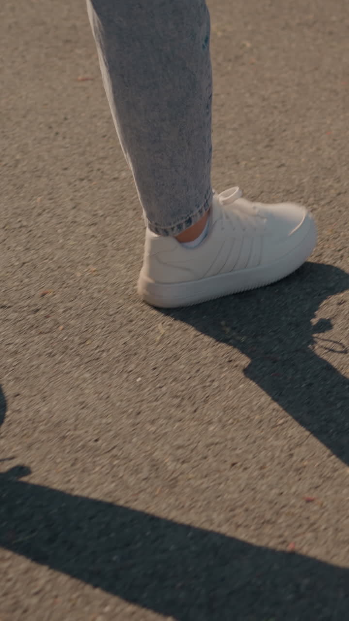 빛이 에 긴 그림자를 던지면서 색 스니커와 청바지를 입은 개인이 타르로 된 도로를 고 있는 측면 시각