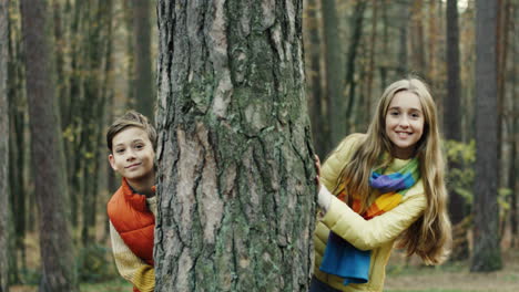 retrato de un chico y una chica lindos y alegres mirando la cámara detrás de un tronco de árbol en el bosque