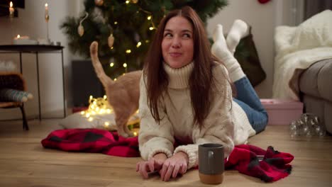 Happy-and-playful-brunette-girl-in-a-White-sweater-plays-with-her-cream-colored-cat-lying-on-a-Red-blanket-in-a-cozy-room-decorated-in-the-New-Year-style