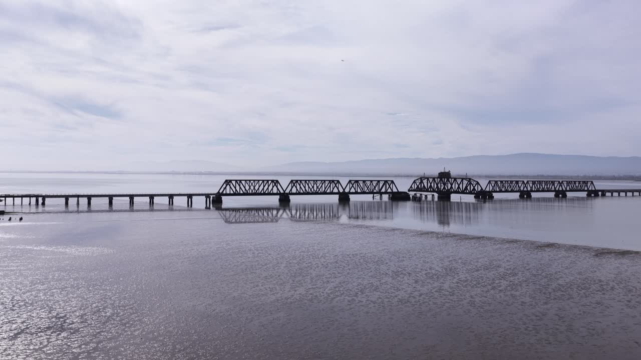 Spettacolari riprese aeree del ponte ferroviario abbandonato di Dumbarton, uno storico ponte girevole che attraversa la Baia di San Francisco, con il suo riflesso visibile sull'acqua calma sotto un cielo nuvoloso