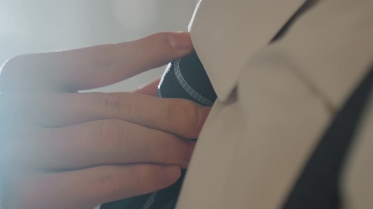 Tangan close-up dengan cahaya lembut sedang merapikan dasi, dasi biru tua bertekstur dengan pola halus dan kerah kemeja yang rapi, suspender terlihat dalam cahaya latar hangat, pengantin pria atau eksekutif yang siap menyelesaikan sentuhan akhir di pagi yang elegan