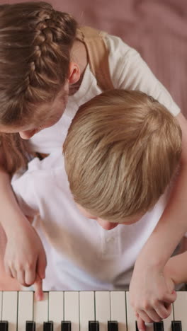 blonde girl holds brother fingers and plays melody on piano in bedroom upper view. adorable little children learn music playing on instrument keyboard at home