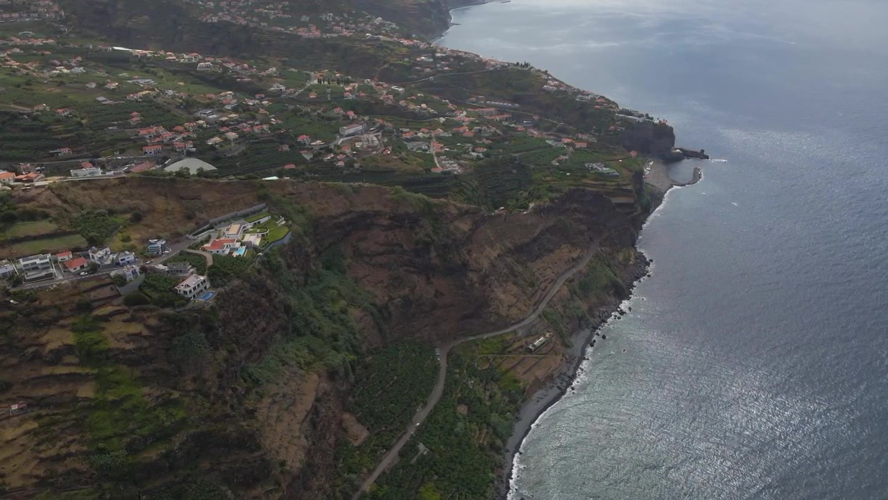 Orbita fluida del drone che cattura le colline a strati di Canhas con l'oceano visibile in lontananza, enfatizzando la drammatica elevazione dalla campagna agricola fino alla costa.