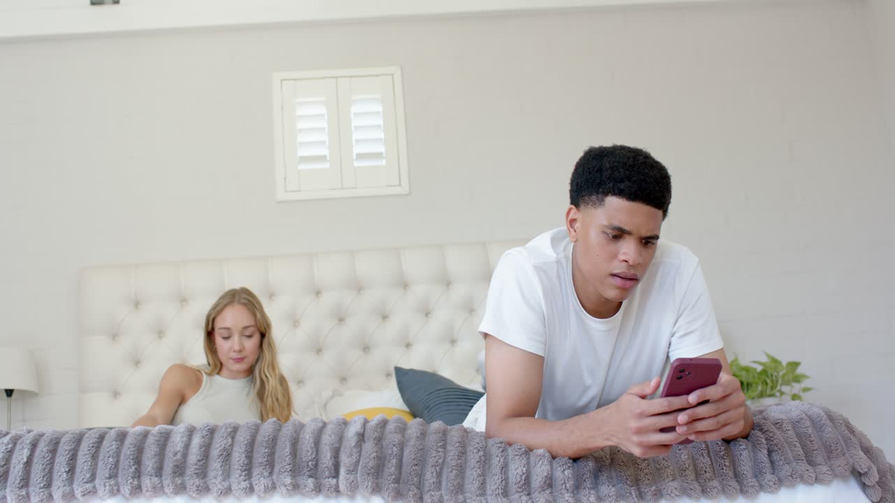 Diverse couple on bed talking after man reading message on pink phone and gesturing, woman watching