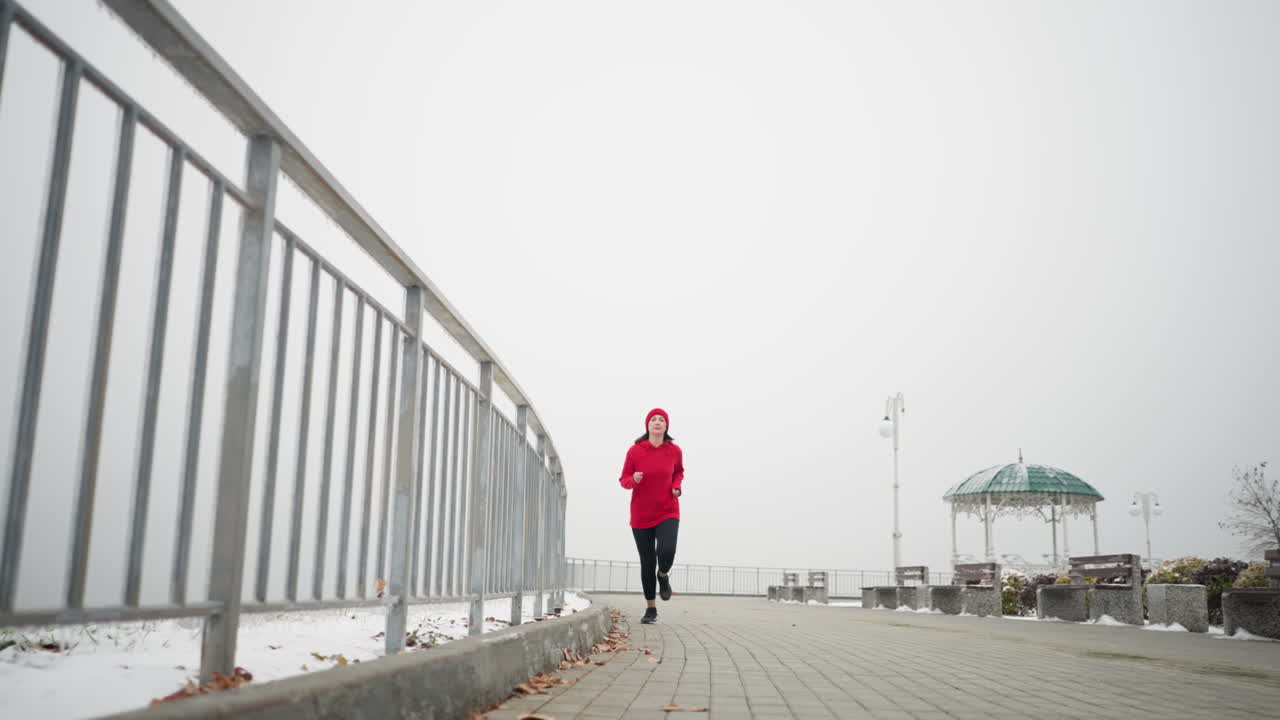 vista de ángulo bajo de una mujer corriendo con gorra roja y chaqueta a lo largo de un camino nevado cerca de una barandilla de hierro, atmósfera brumosa con estructura decorativa y bancos en el fondo