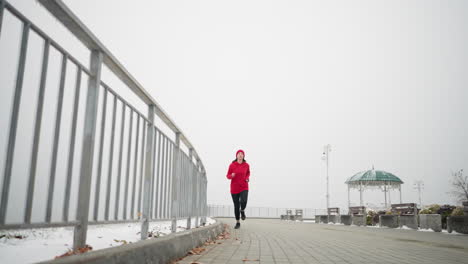 vista de ángulo bajo de una mujer corriendo con gorra roja y chaqueta a lo largo de un camino nevado cerca de una barandilla de hierro, atmósfera brumosa con estructura decorativa y bancos en el fondo