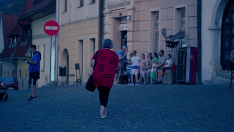 Rückaufnahme-Einer-älteren-Weißen-Frau-Mit-Rotem-T-Shirt,-Die-Im-Sommer-Nachts-In-Zeitlupe-Durch-Die-Historische-Stadt-Spaziert,-Mit-Wunderschönem-Bokeh-Aus-Orangefarbenen-Lichtern-Im-Hintergrund