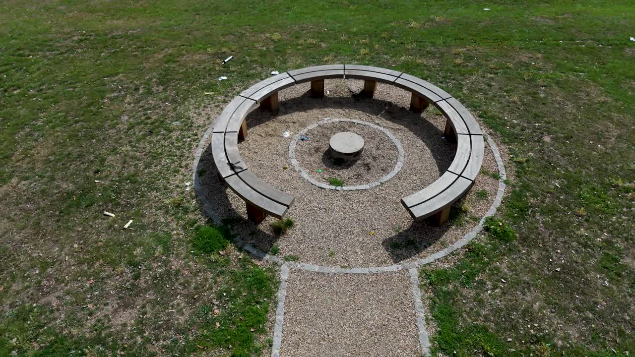 Low drone flyover of circular wooden bench with white lines and stool in a park