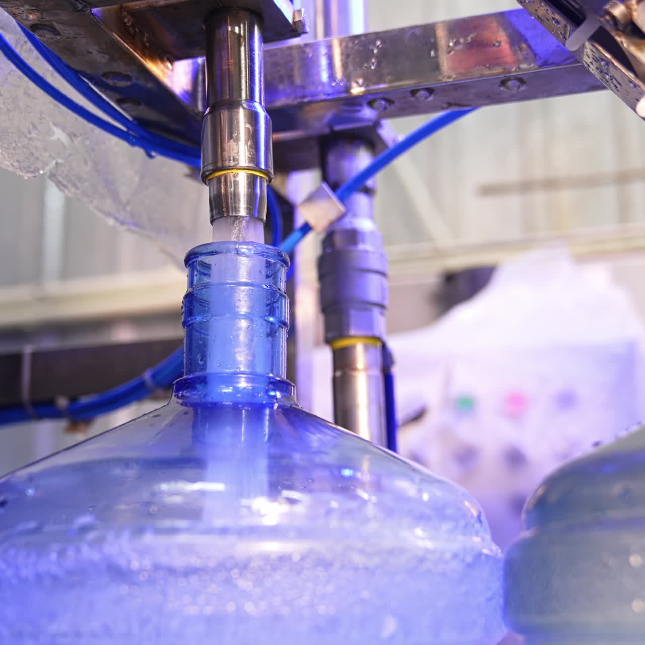 Clean water is filling the big blue bottle from a metal tube. Water overflows the flask. Close up